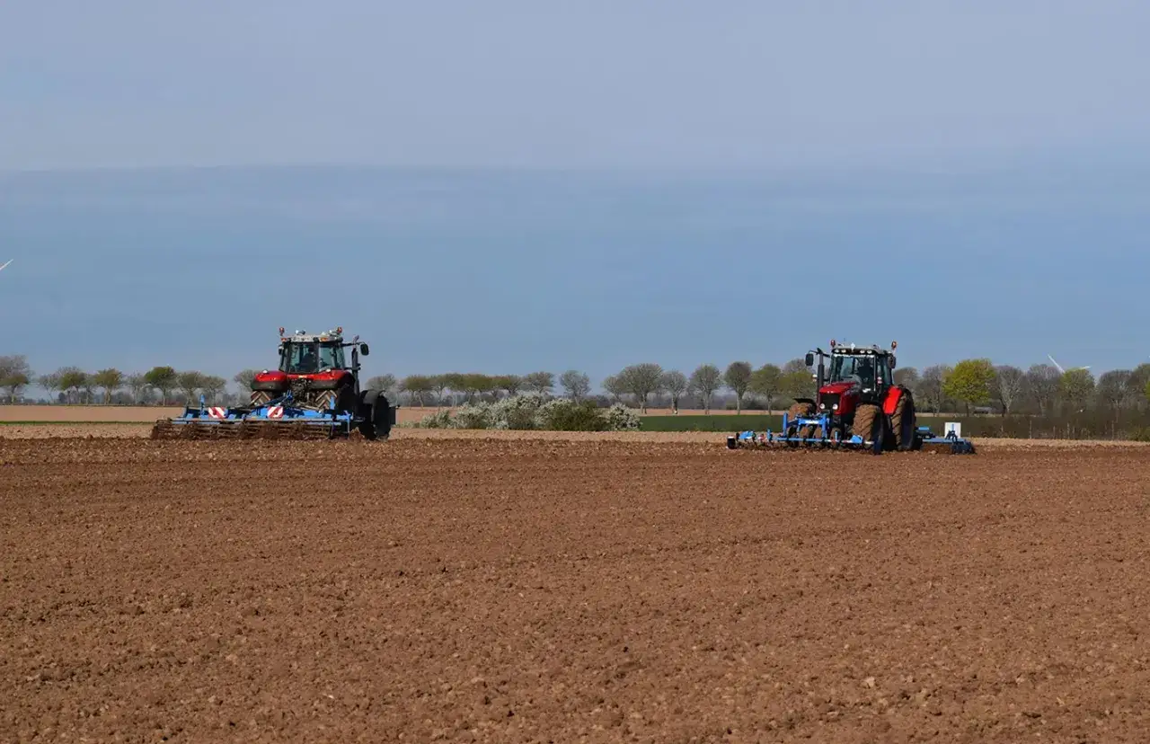 Deux tracteurs rouges travaillent dans un champ labouré, préparant le sol pour la culture du lin.