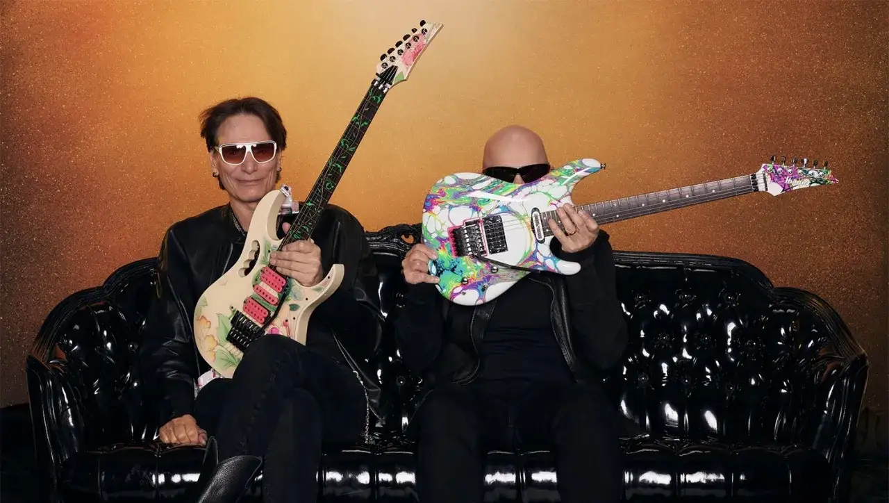 Steve Vai i Joe Satriani, dwaj mistrzowie gitary, siedzą na czarnej kanapie z kolorowymi gitarami.