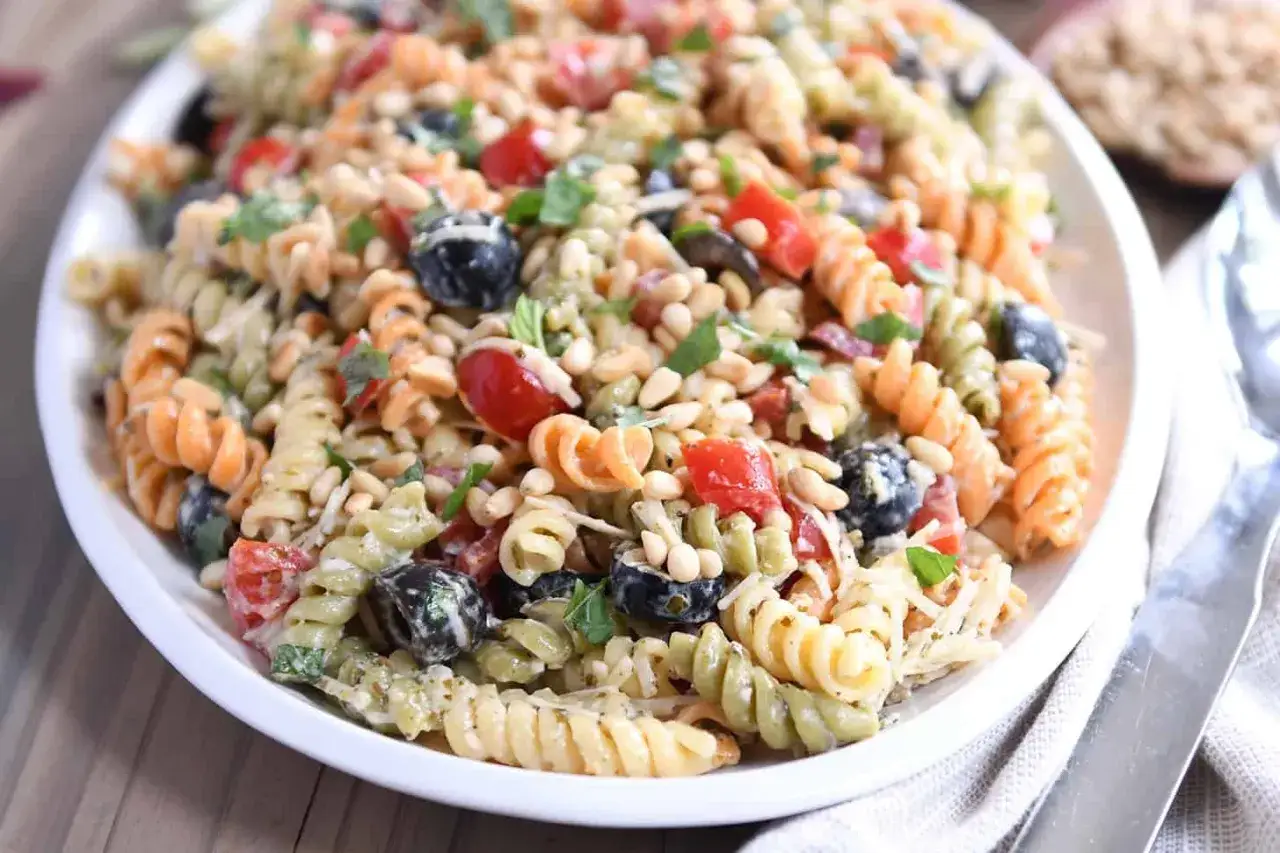 Bunter italienischer Nudelsalat mit Spiralnudeln, Kirschtomaten, schwarzen Oliven und Pinienkernen, serviert auf einem weißen Teller.