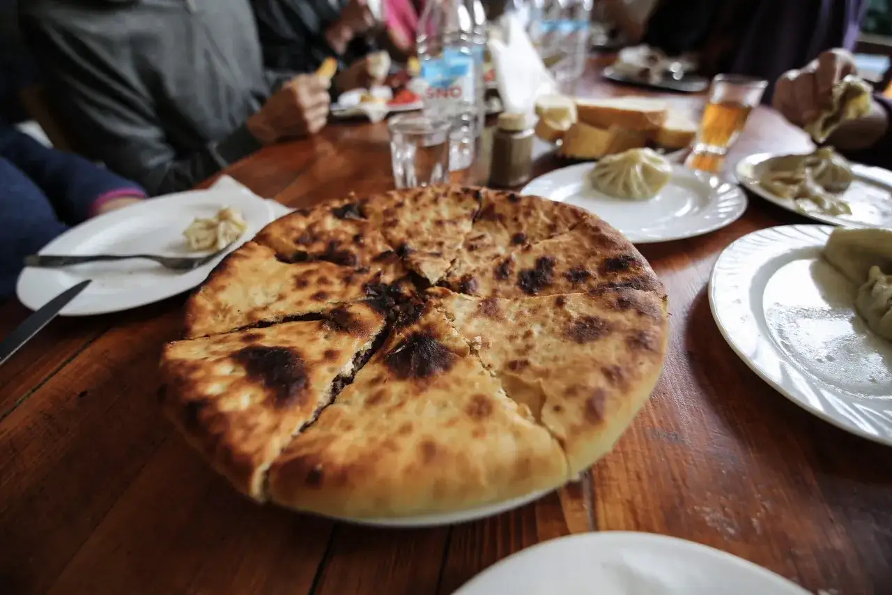 En stor, gyllene khachapuri, en klassiker inom georgisk mat, delas upp p&aring; ett tr&auml;bord. Runtom syns khinkali och andra l&auml;ckerheter.