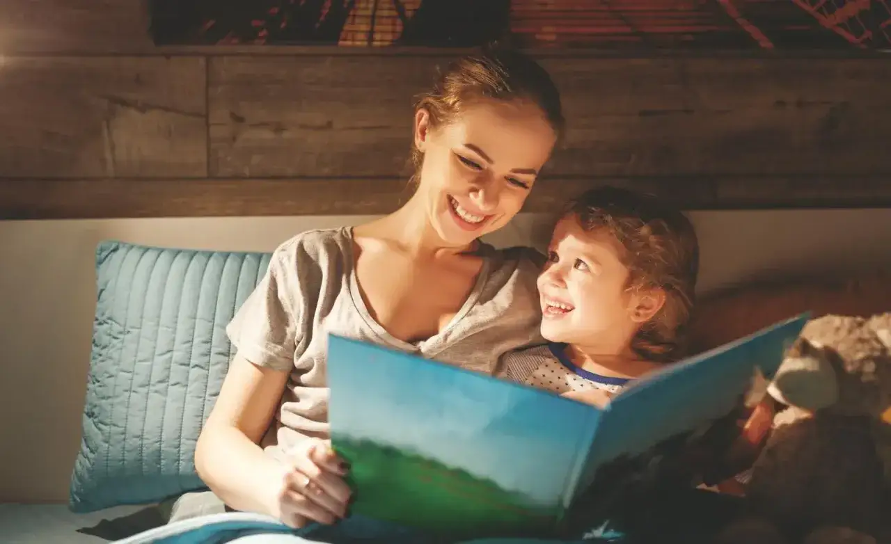 Mama czyta bajkę dziecku, kt&oacute;re ma problemy z czytaniem. Uśmiechnięci patrzą na książkę.