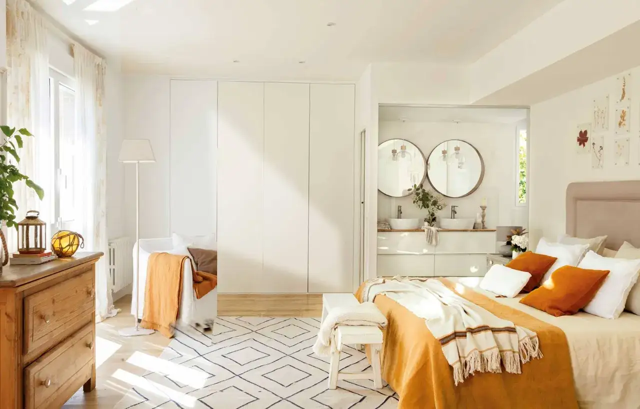 Acogedora habitaci&oacute;n blanca con cama naranja, c&oacute;moda de madera y ba&ntilde;o con lavabos redondos.