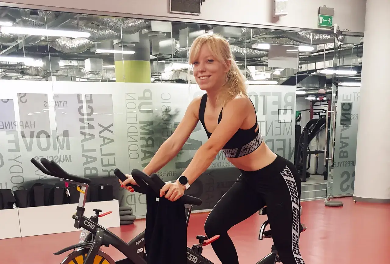 Uśmiechnięta blondynka na rowerze stacjonarnym. Czy indoor cycling to dobry trening? Tak, to świetna forma cardio!