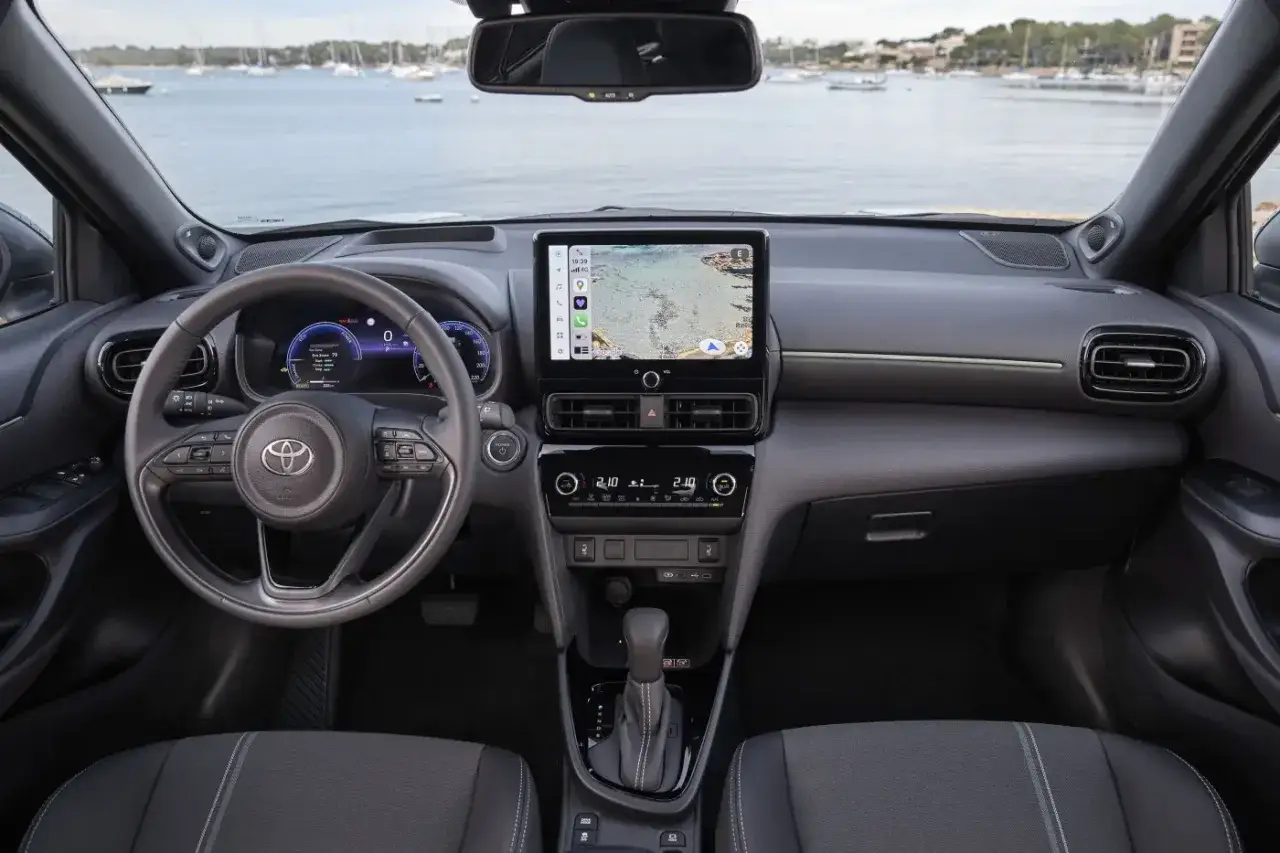 Wnętrze Toyota Yaris Hybrid z ekranem nawigacji i cyfrowymi zegarami. Widok na morze i łodzie przez przednią szybę.