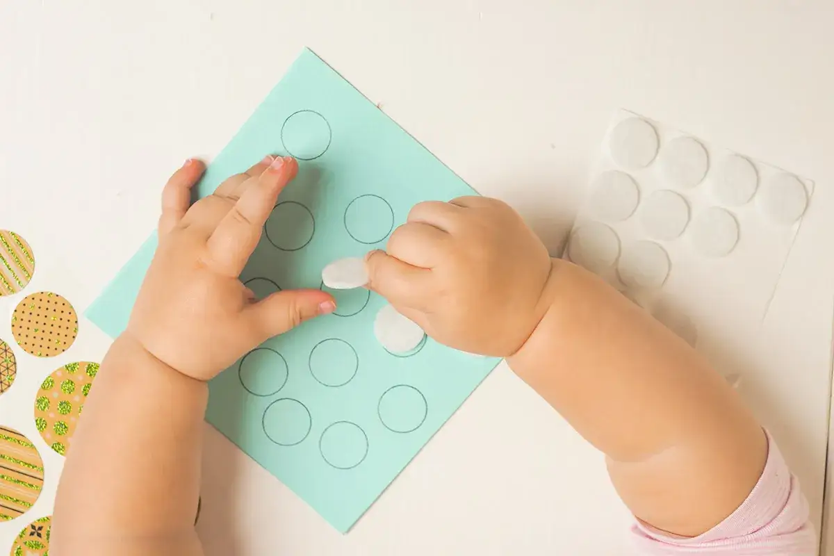 Dziecko tworzy zabawki montessori diy, przyklejając białe kółka na niebieskiej kartce z narysowanymi okręgami.