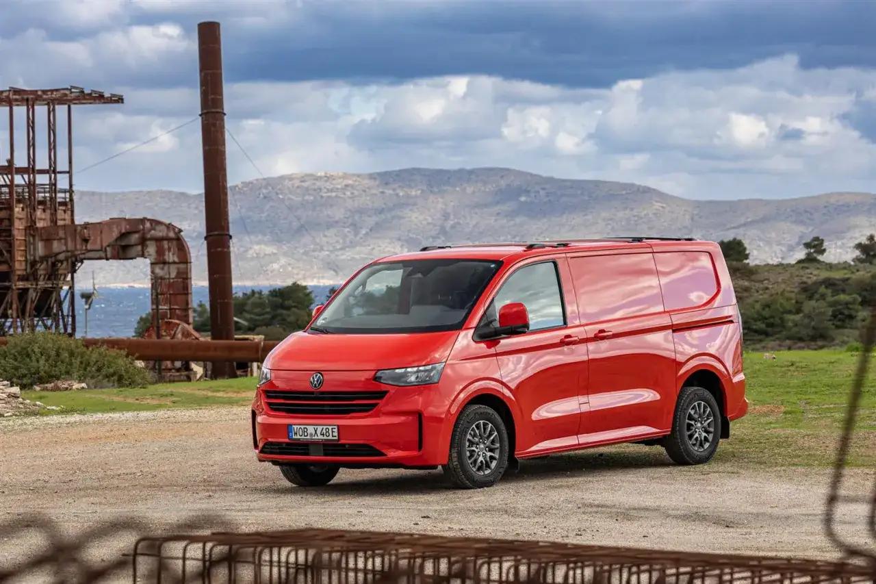 Un furgone rosso Volkswagen Transporter, con le sue dimensioni ideali per ogni avventura, parcheggiato in un paesaggio industriale abbandonato con montagne sullo sfondo.
