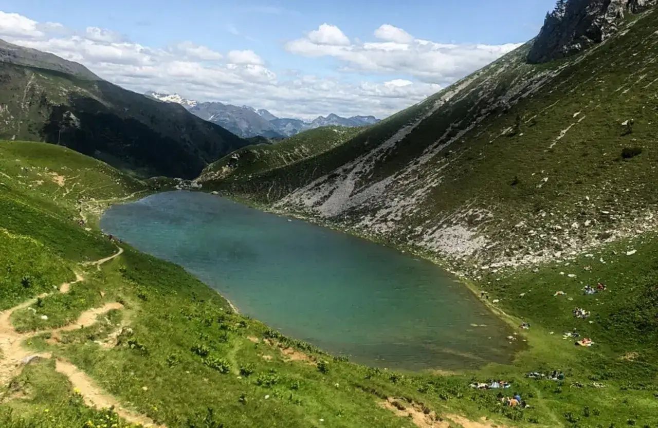Un sentiero serpeggia tra prati verdi verso il lago Branchino, le cui acque turchesi riflettono le montagne circostanti. Alcune persone si godono un picnic sulle rive.