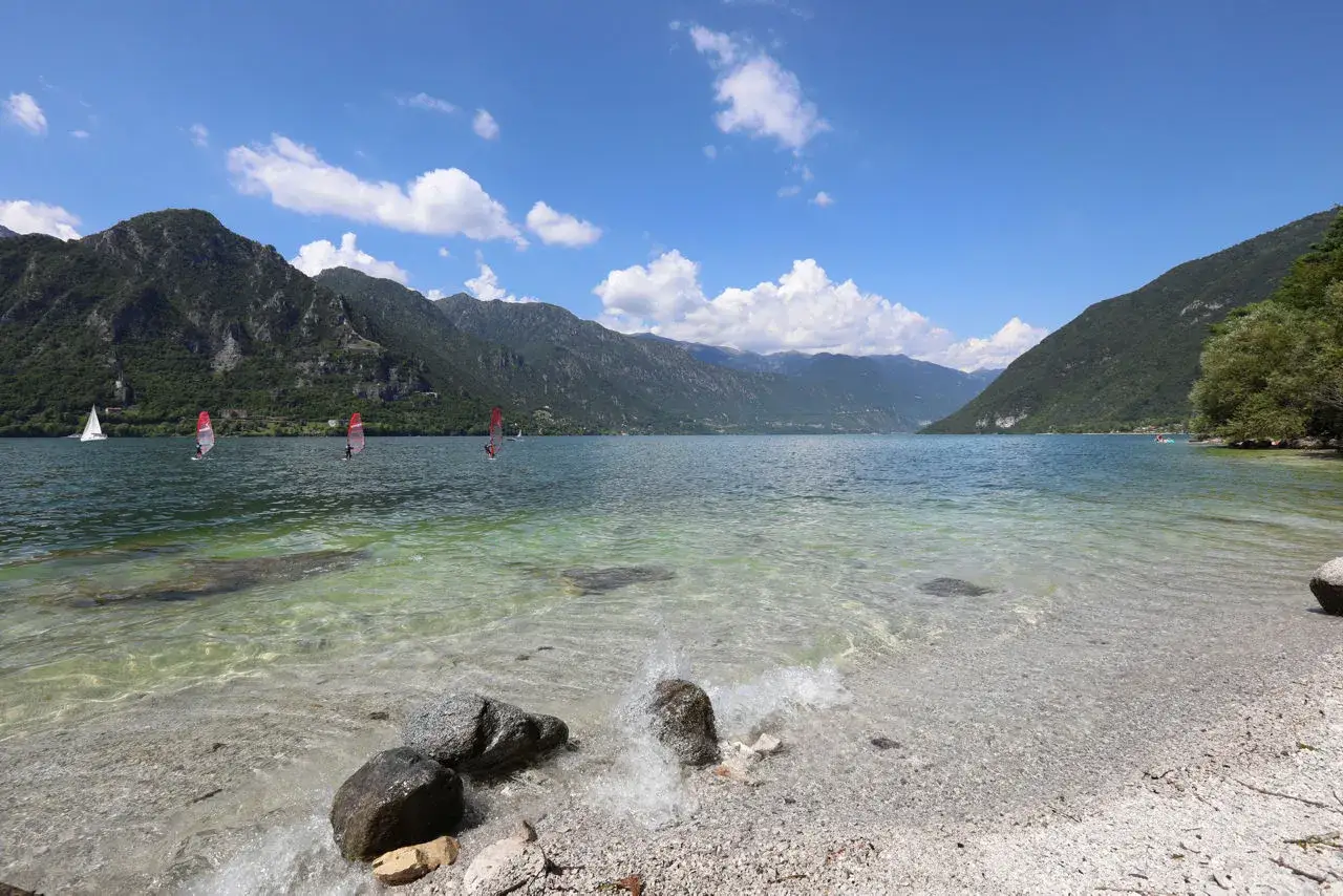 Splendida giornata al lago vicino Milano con spiaggia di ciottoli. Veleggiatori e windsurfisti solcano le acque limpide, circondati da montagne verdi.