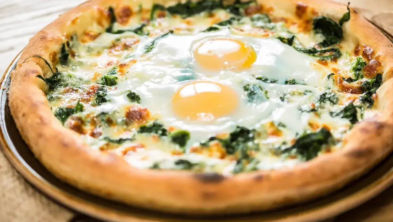Pyszna pizza z jajkiem i szpinakiem, z dwoma idealnie wpasowanymi jajkami na wierzchu, otoczona puszystym ciastem.
