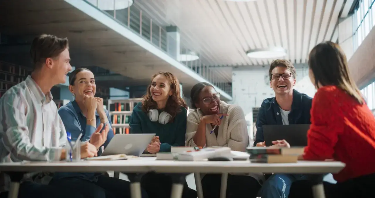 Lachende Studenten im Studium, vielleicht auf dem Weg zum Master Gesundheitspsychologie, tauschen sich an einem Tisch aus.