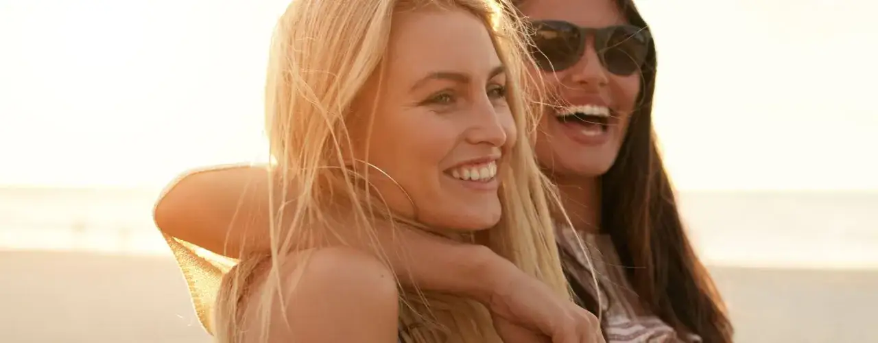 Zwei lachende Freundinnen am Strand. Die Blonde fragt sich, wie werde ich braun mit heller Haut, während die Dunkelhaarige die Sonne genießt.