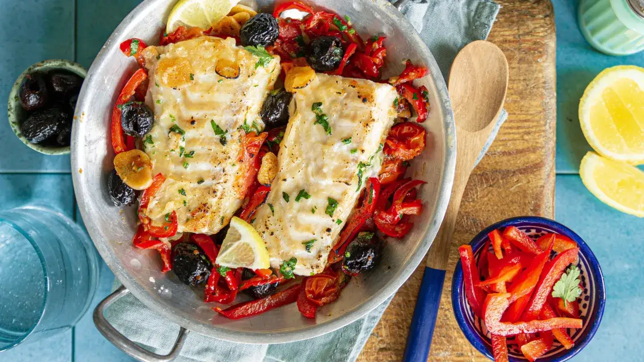Deux beaux dos de cabillaud cuits avec des poivrons rouges, des olives noires et des quartiers de citron dans un plat en métal.