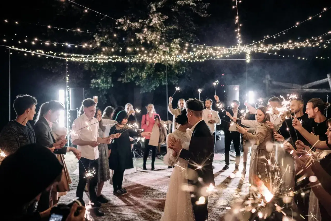 Un couple danse sous des guirlandes lumineuses, entour&eacute; d'invit&eacute;s tenant des feux de Bengale. L'ambiance parfaite pour une playlist mariage pour danser.
