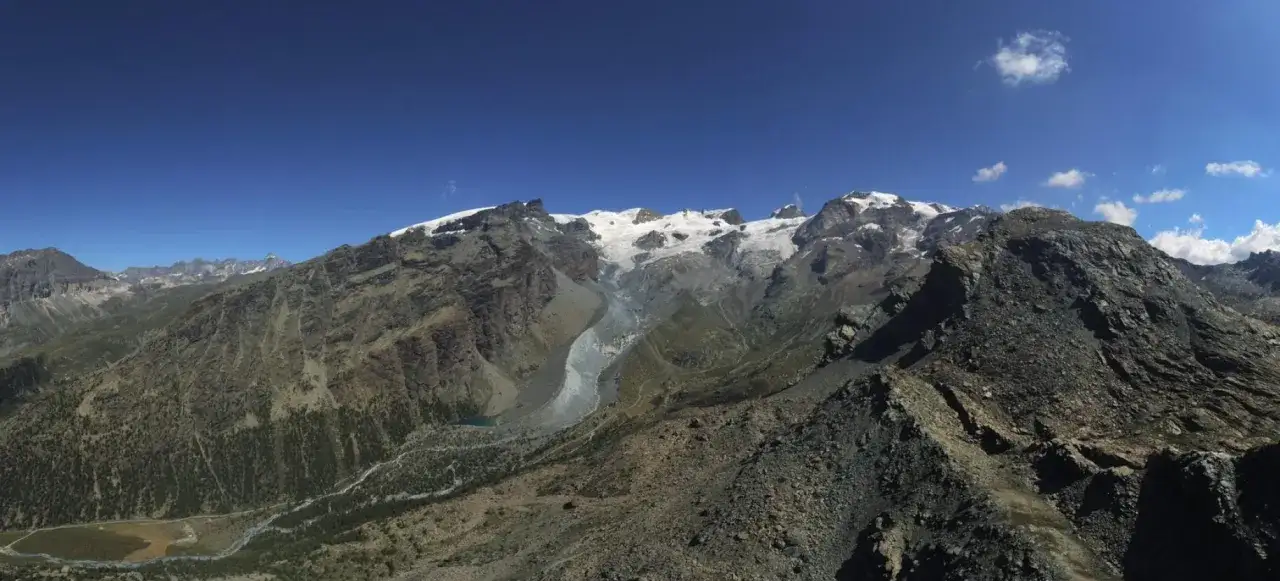 Vasto panorama alpino con cime innevate, un ghiacciaio che scende a valle e un torrente che serpeggia tra rocce e vegetazione. Il **palon di Resy** domina il paesaggio.