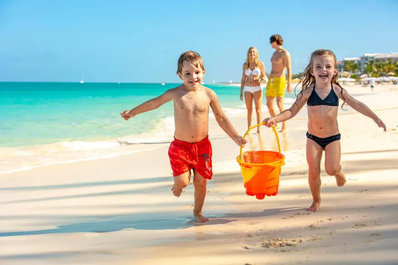 Radosne dzieci na plaży biegną z wiaderkiem, a w tle widać rodziców i turkusowe morze.