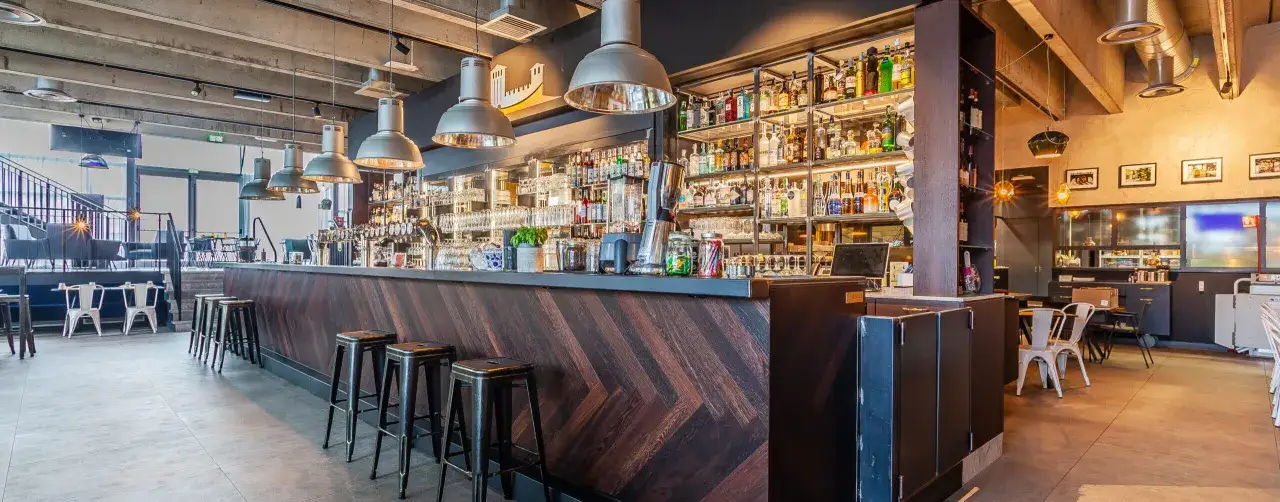 Interno di un locale moderno con bancone in legno a spina di pesce, sgabelli alti e scaffali pieni di bottiglie. Un'atmosfera accogliente che ricorda un birrificio Legnano.