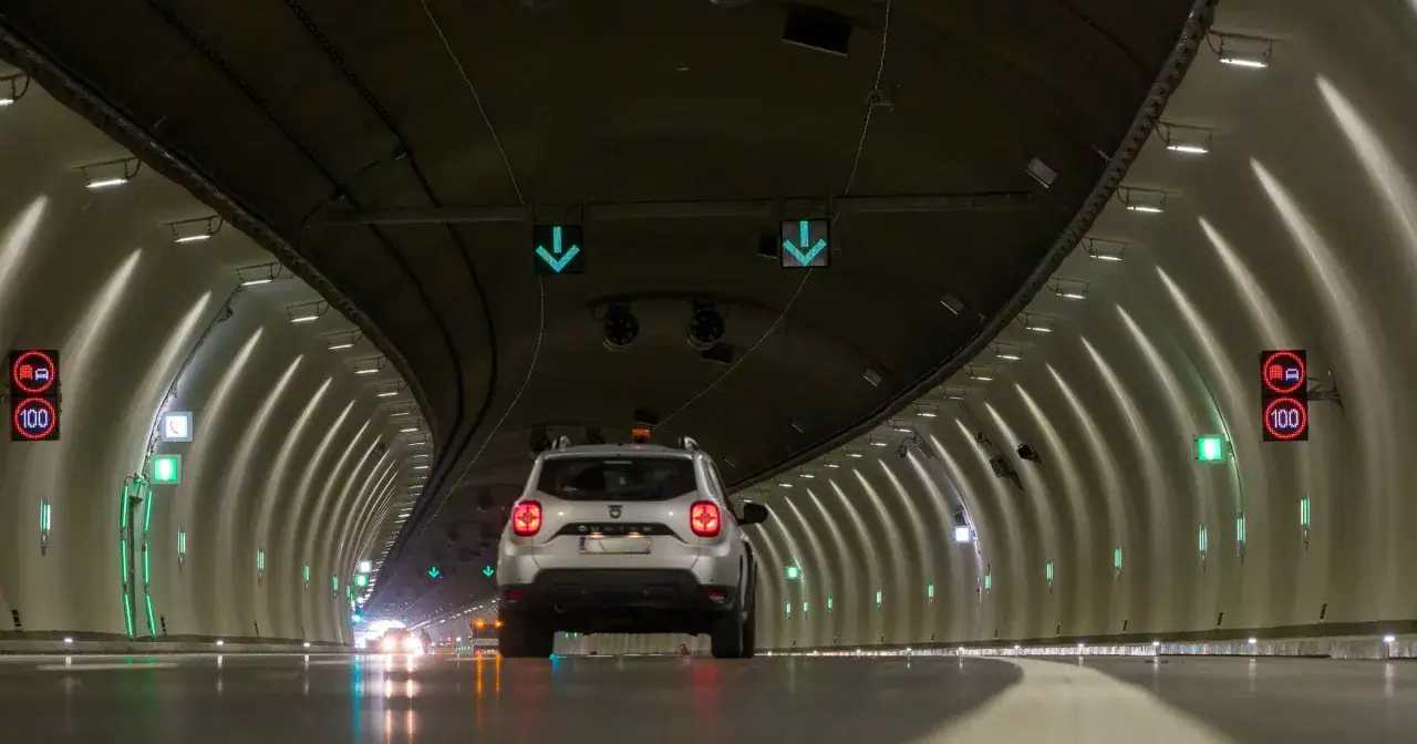 Srebrny SUV jedzie przez nowoczesny tunel na Zakopiance. Zielone strzałki i znaki ograniczenia prędkości 100 km/h oświetlają drogę.