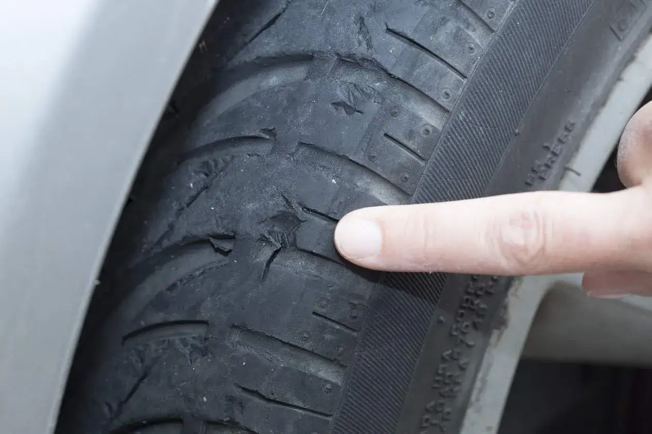 Dedo señalando el desgaste irregular de neumáticos, con grietas y trozos faltantes en la banda de rodadura.