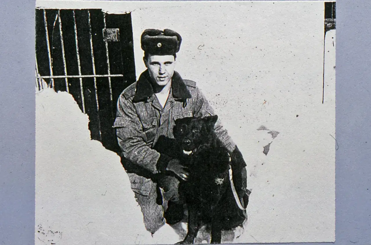 Ein junger Mann, Dietmar Schultke, in Uniform und mit M&uuml;tze, sitzt im Schnee und h&auml;lt einen schwarzen Hund.
