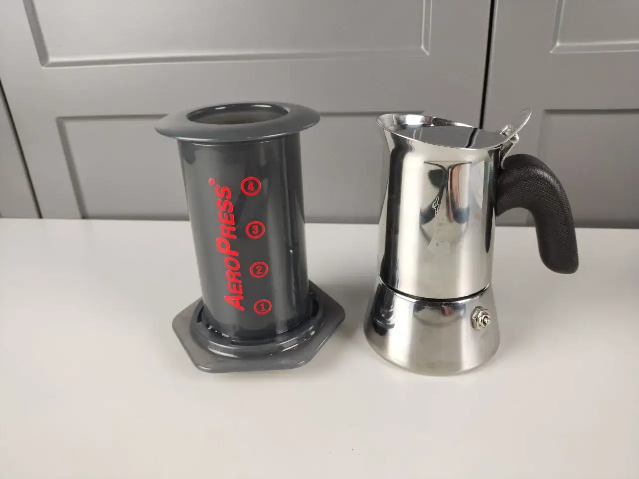 Aeropress i błyszcząca kawiarka stoją obok siebie, gotowe do parzenia kawy. Które wybierzesz?