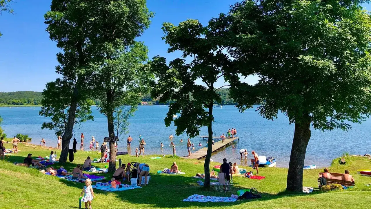 Słoneczna plaża nad jeziorem tętni życiem. Ludzie odpoczywają na trawie, kąpią się w wodzie i spacerują po molo.