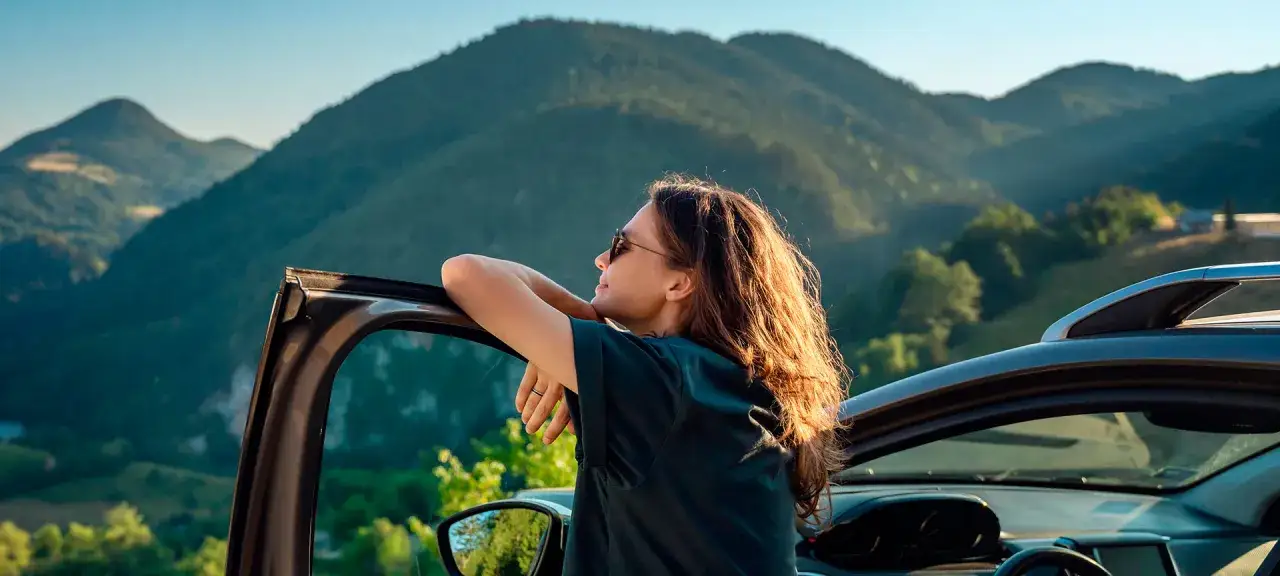 Mujer con gafas de sol apoyada en la puerta de un coche, disfrutando de un paisaje monta&ntilde;oso. El viaje en coche le permite contemplar la belleza natural.