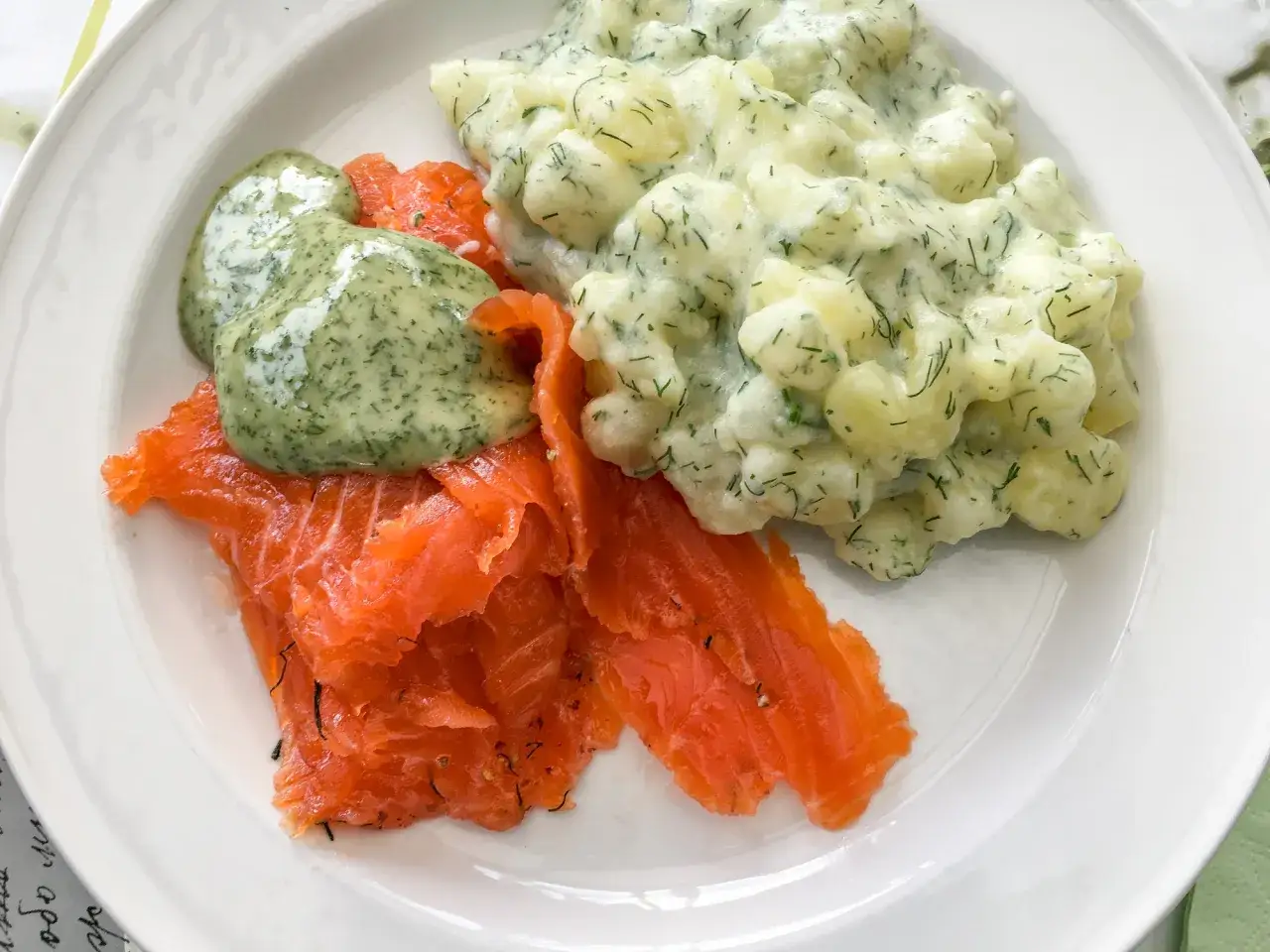 En tallrik med gravad lax, potatissallad med dill och en grön sås.