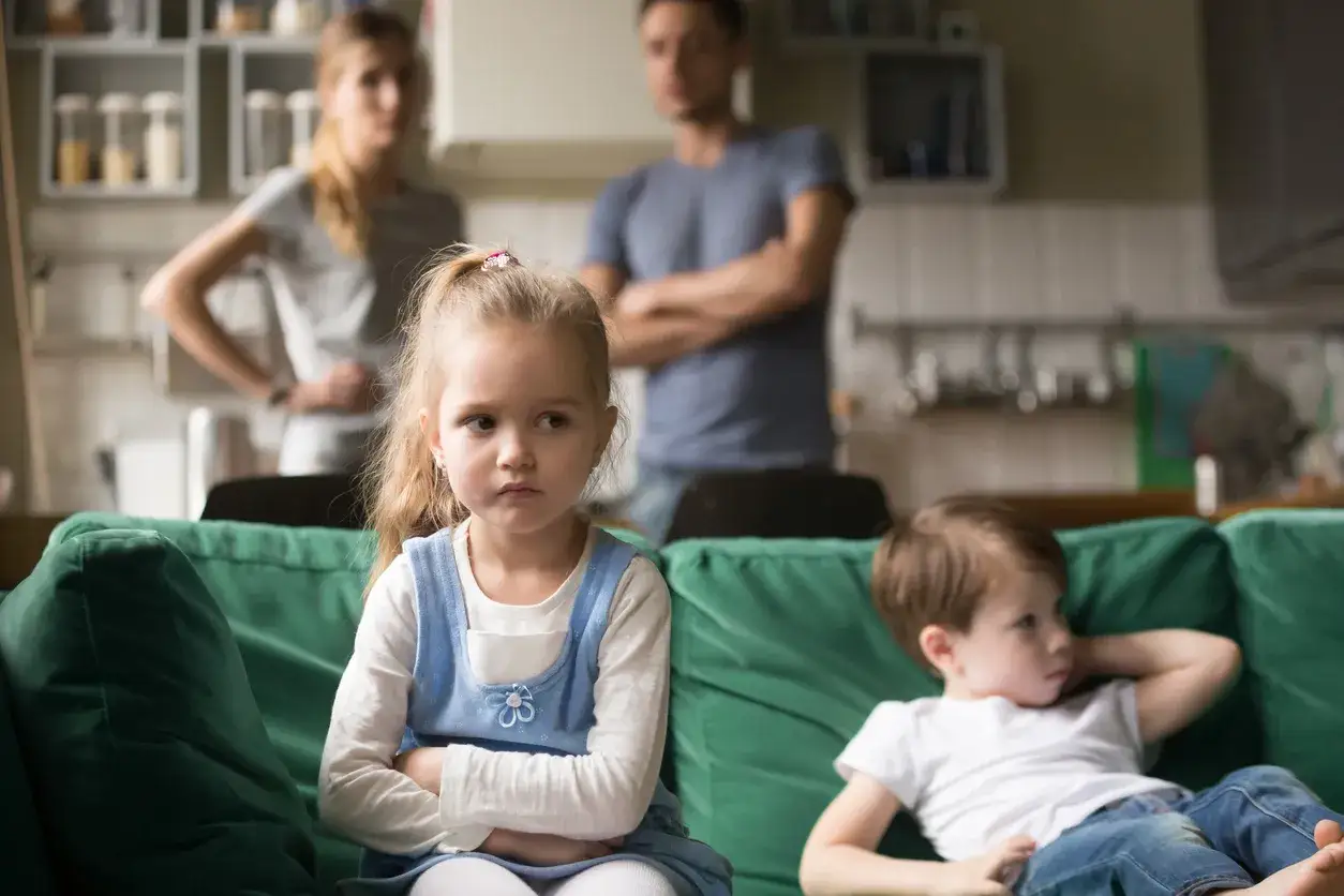Zazdrość u dzieci w wieku szkolnym. Dziewczynka z obrażoną miną siedzi na kanapie, obok niej chłopiec. Rodzice stoją w tle.