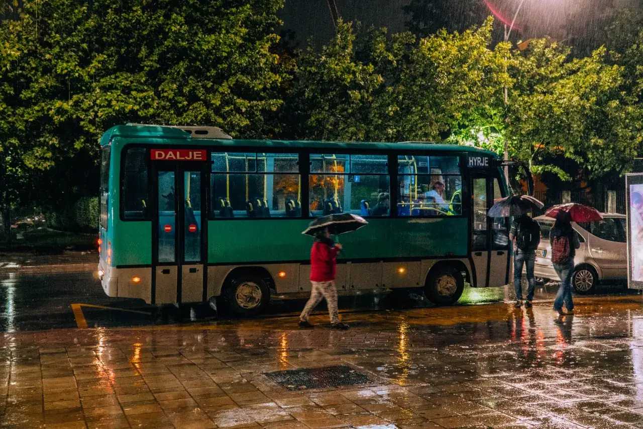 Deszczowy wiecz&oacute;r w Albanii. Ludzie z parasolami czekają na autobus, odbijając się w mokrej nawierzchni.