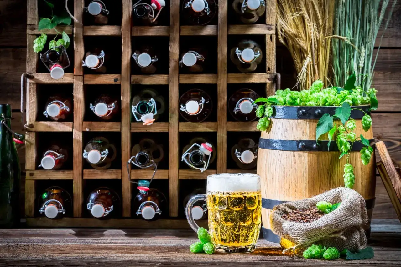 Piwniczka z piwem craftowym: butelki w drewnianym stojaku, beczka z chmielem, kufel złocistego trunku. Jak przechowywać piwo craftowe w pionie czy w poziomie?