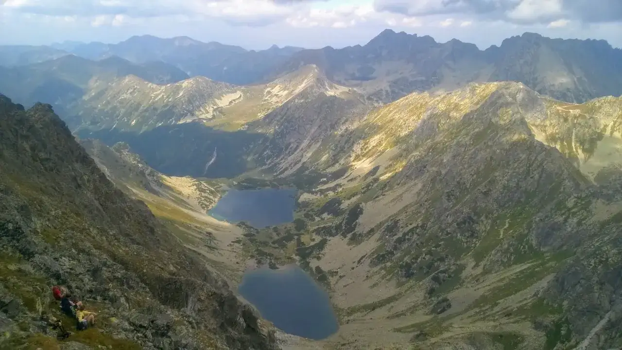 Widok na dwa górskie jeziora w **Tatrach Słowackich**. Na pierwszym planie grupa turystów odpoczywa na skalistym zboczu.