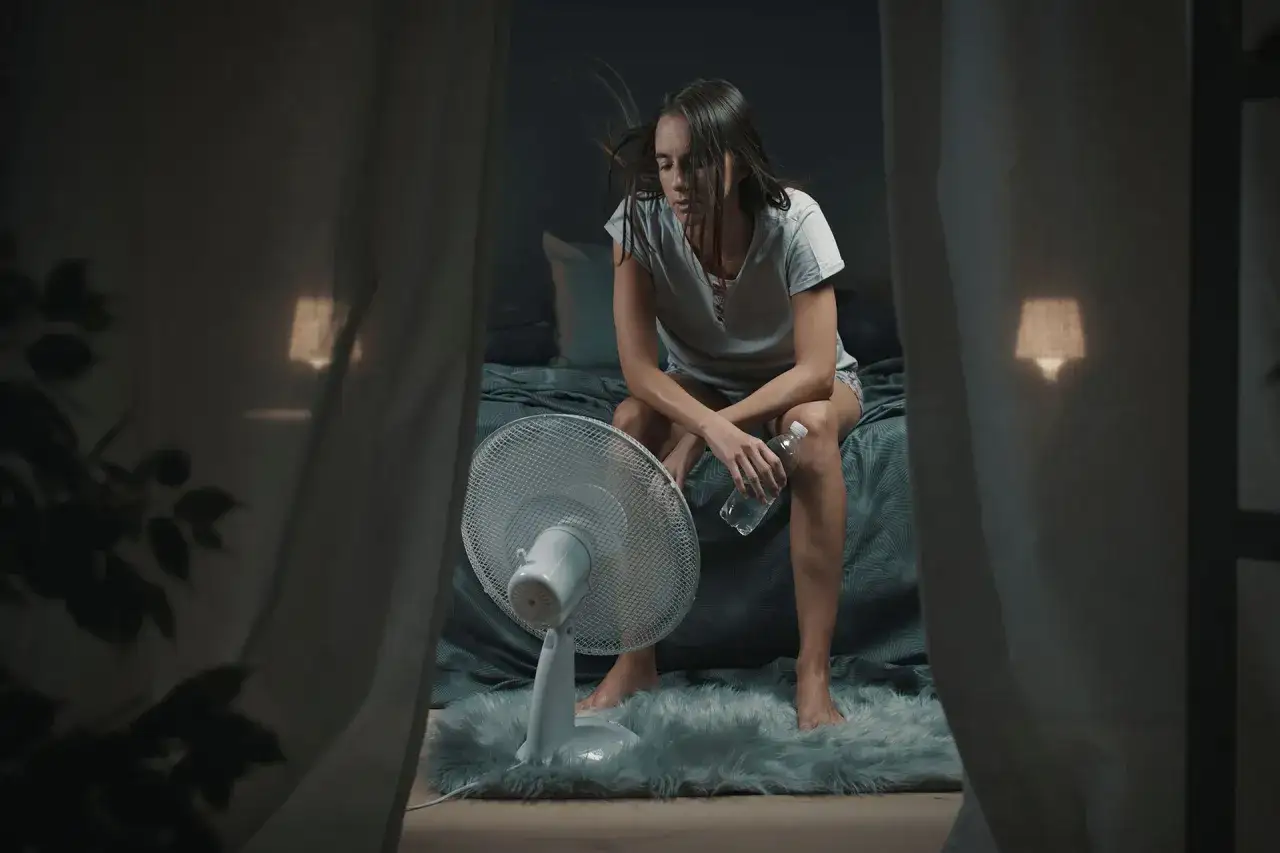 Donna seduta sul letto, con un ventilatore acceso, lotta contro il caldo soffocante e la depressione. L'aria immobile amplifica il suo disagio.