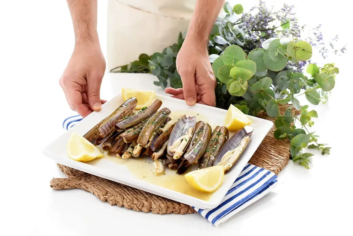 Manos sirviendo un plato de navajas animal, con limón y hierbas, decorado con eucalipto y flores moradas.