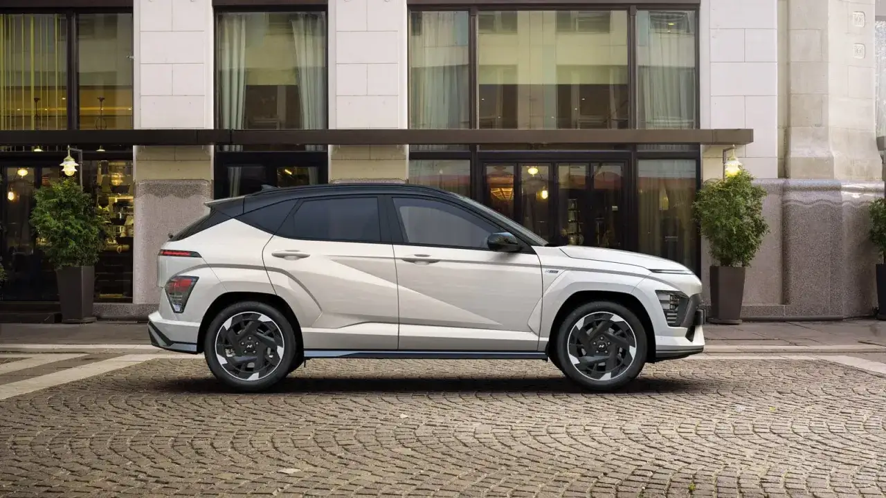 Nowy Hyundai Kona w kolorze jasnoszarym, zaparkowany na brukowanej ulicy przed budynkiem. Sprawdź hyundai kona cena!