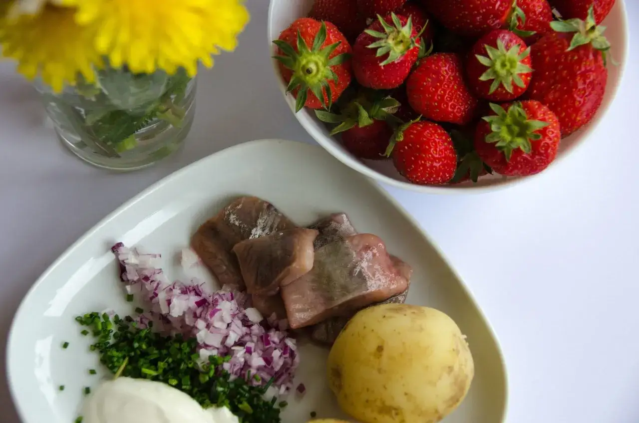 En tallrik med sill, potatis, gräslök, rödlök och gräddfil. En skål med jordgubbar och en bukett med gula blommor. En försmak på vad man har på midsommarbordet.