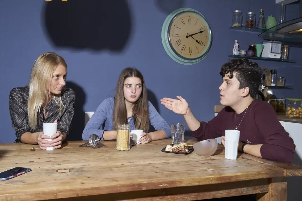 Ein Junge gestikuliert wild, während seine Mutter und Schwester ihn am Tisch ansehen. Ein deutlicher **Streit in der Familie** liegt in der Luft.
