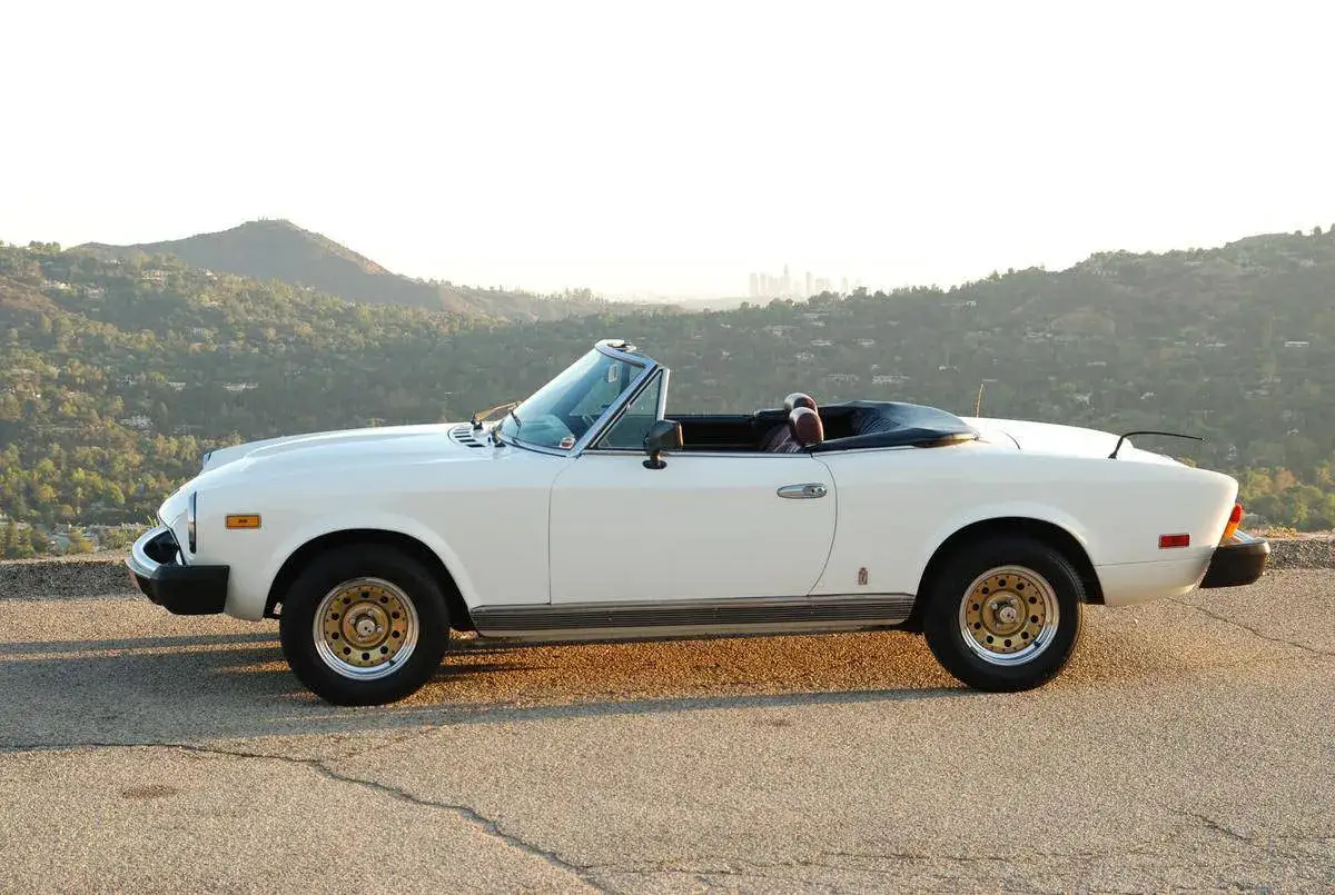 Biały kabriolet Fiat X1/9 z lat 70. na tle wzgórz i miasta.
