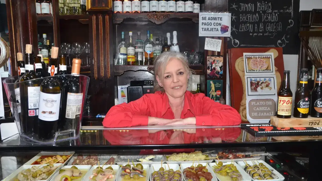 Una mujer sonríe detrás de la barra de salsipuedes una tasca encantadora, con botellas de vino y tapas expuestas.