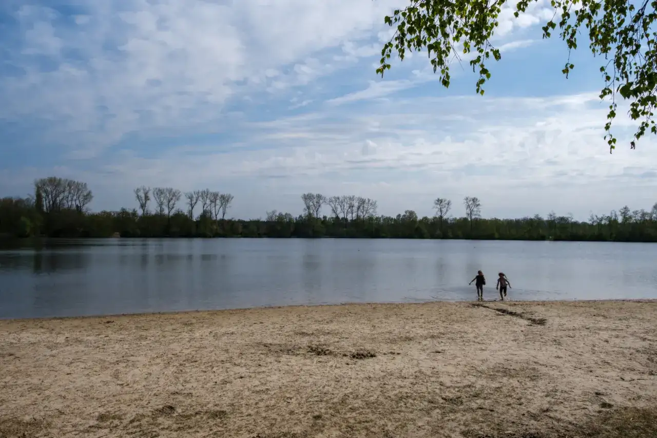 Zwei Kinder planschen im flachen Wasser am Sandstrand des Dieksees in Lingen. B&auml;ume s&auml;umen das Ufer unter einem bew&ouml;lkten Himmel.