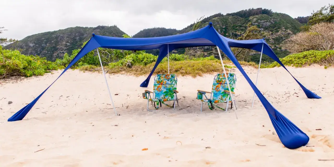 Tv&aring; strandstolar med tropiskt m&ouml;nster st&aring;r under ett bl&aring;tt solskydd p&aring; en sandstrand. Perfekt f&ouml;r att njuta av utsikten, &auml;ven om man inte vill att blir man brun i skuggan.