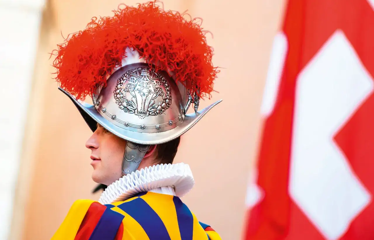 Gwardia szwajcarska w historycznym stroju, z ozdobnym hełmem i pióropuszem, stoi na tle flagi Watykanu.