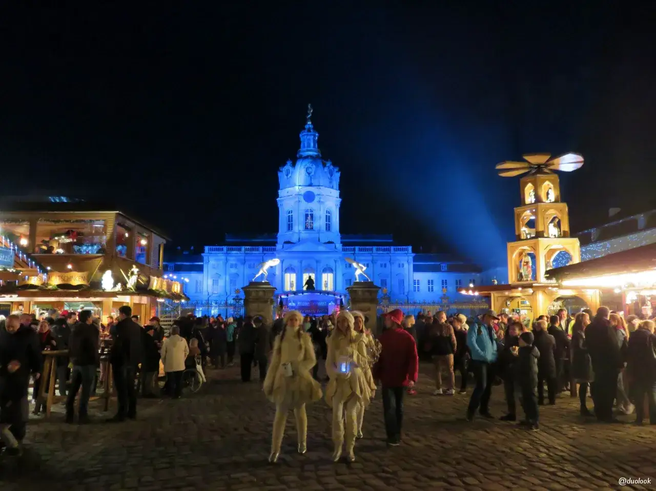 Nocny jarmark w Berlinie, z oświetlonym pałacem w tle i ludźmi spacerującymi między straganami.