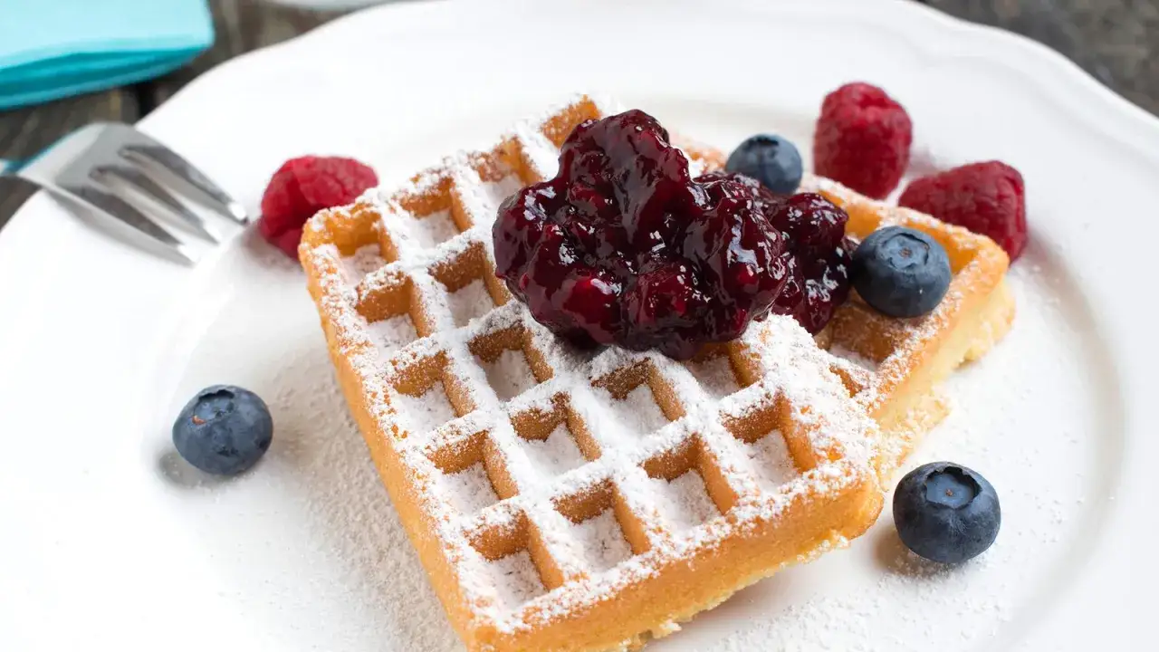 Zwei Buttermilch Waffeln mit Puderzucker, Beeren und Marmelade auf einem weißen Teller.