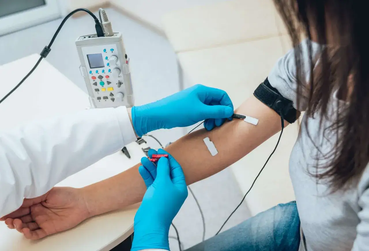 Lekarz w niebieskich rękawiczkach przeprowadza próbę tężyczkową, podłączając elektrody do ramienia pacjentki.