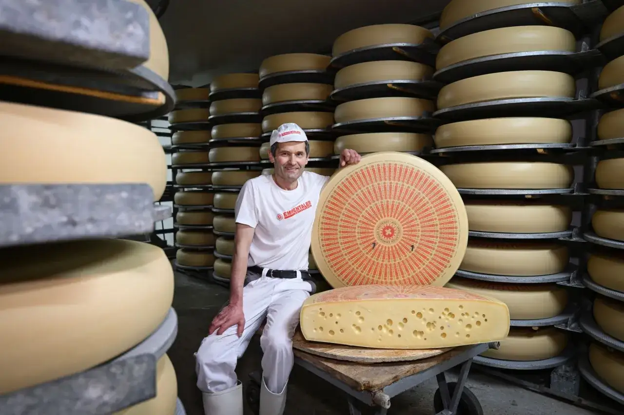 Ein Mann in wei&szlig;er Kleidung posiert stolz neben einem riesigen Laib Emmentaler K&auml;se. Die vielen runden K&auml;selaibe im Hintergrund zeugen von der reichen Emmentaler Herkunft.