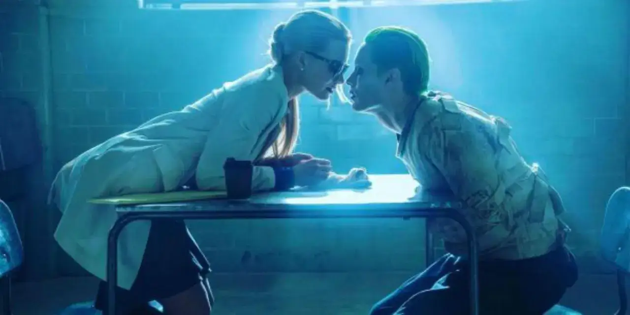 Joker i Harley pochylają się nad stołem, ich twarze blisko siebie. Harley w okularach i białym płaszczu, Joker z zielonymi włosami.