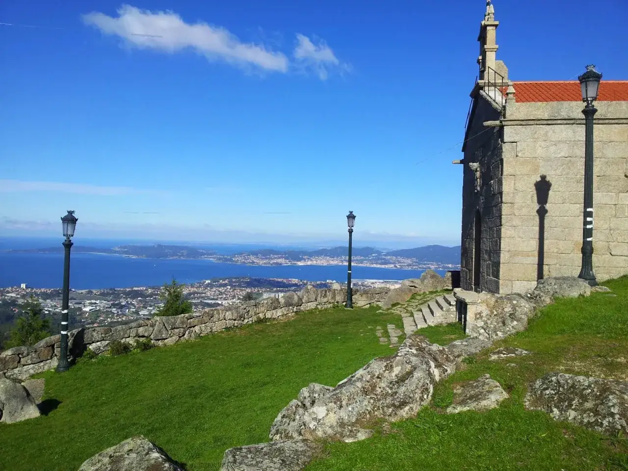 Vista desde el monte Alba: una ermita de piedra con tejado rojo, farolas y un mar azul al fondo con una ciudad costera.