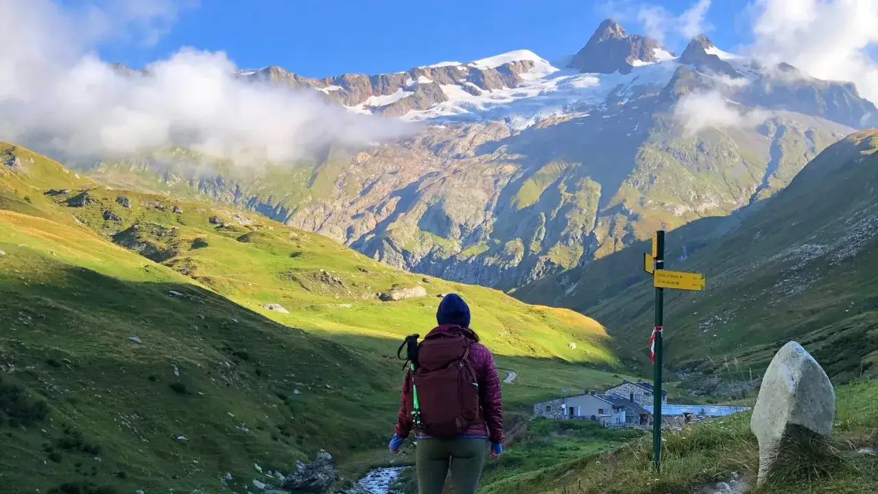 A hiker pauses on a **Tour Mont Blanc hike**, gazing at a majestic snow-capped mountain range. A signpost points the way to mountain huts.