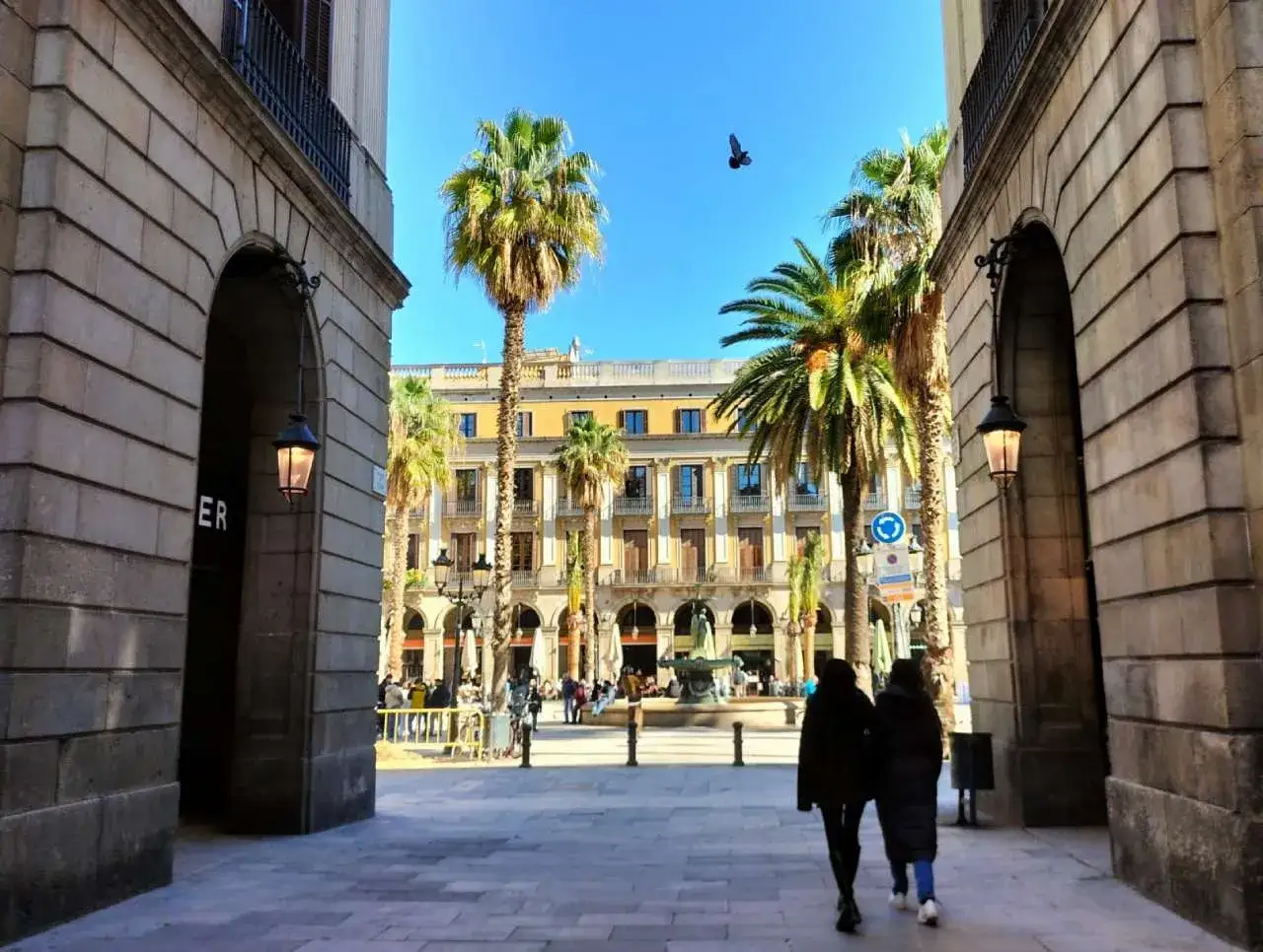 Zwei Personen spazieren durch eine Gasse mit Palmen und historischen Geb&auml;uden in Barcelona im April.
