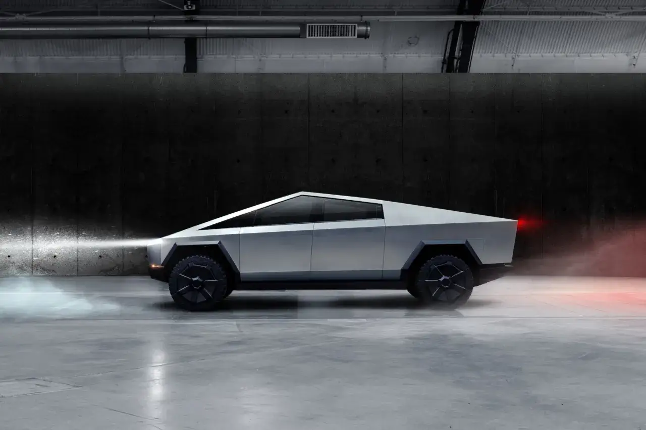 Futurystyczny Tesla Cybertruck z ostrymi liniami i masywnymi kołami, oświetlony reflektorami i czerwonymi światłami pozycyjnymi, w industrialnym wnętrzu.